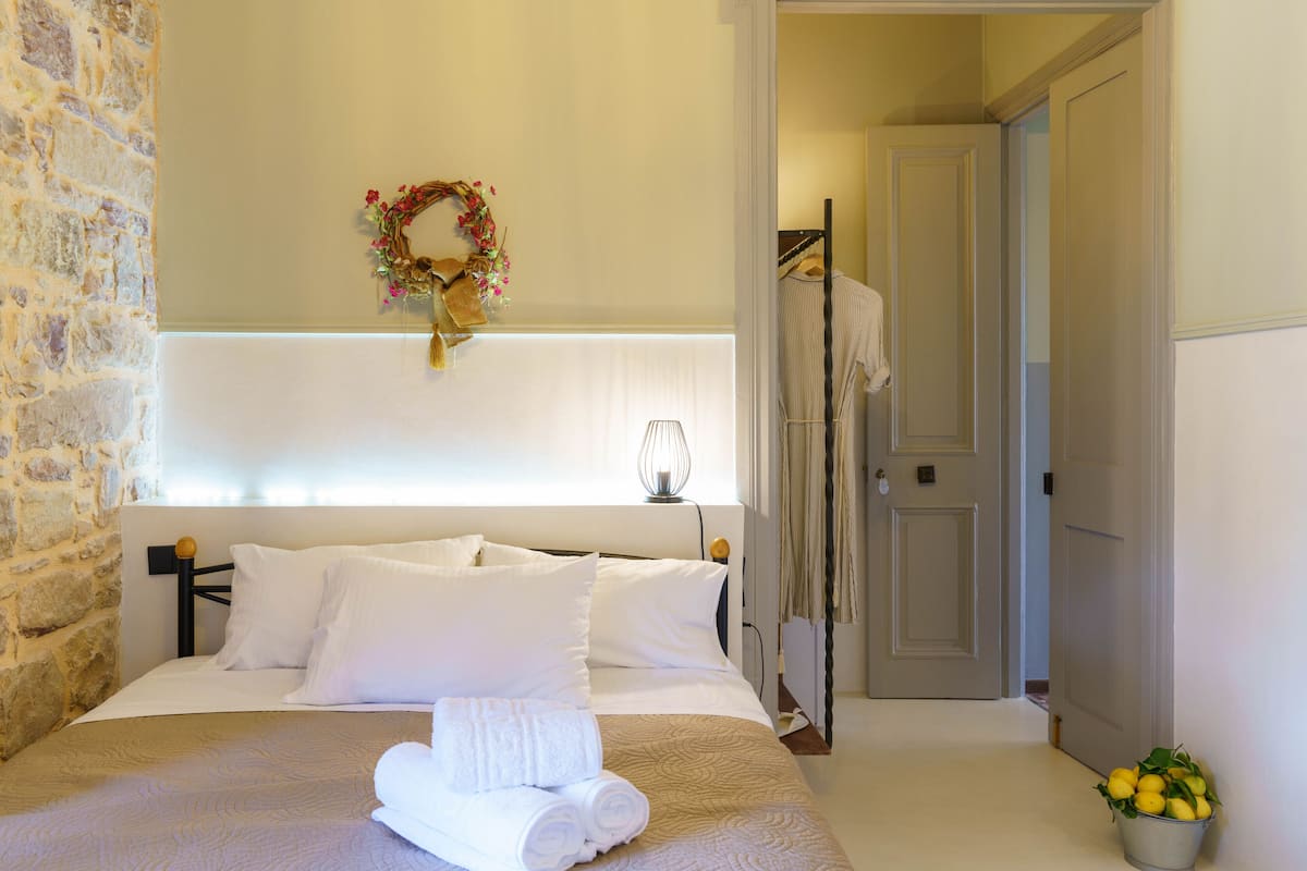 Una cama bien hecha con sábanas blancas y una toalla enrollada, una lámpara fijada a la pared y un pequeño cubo de limones en el suelo.