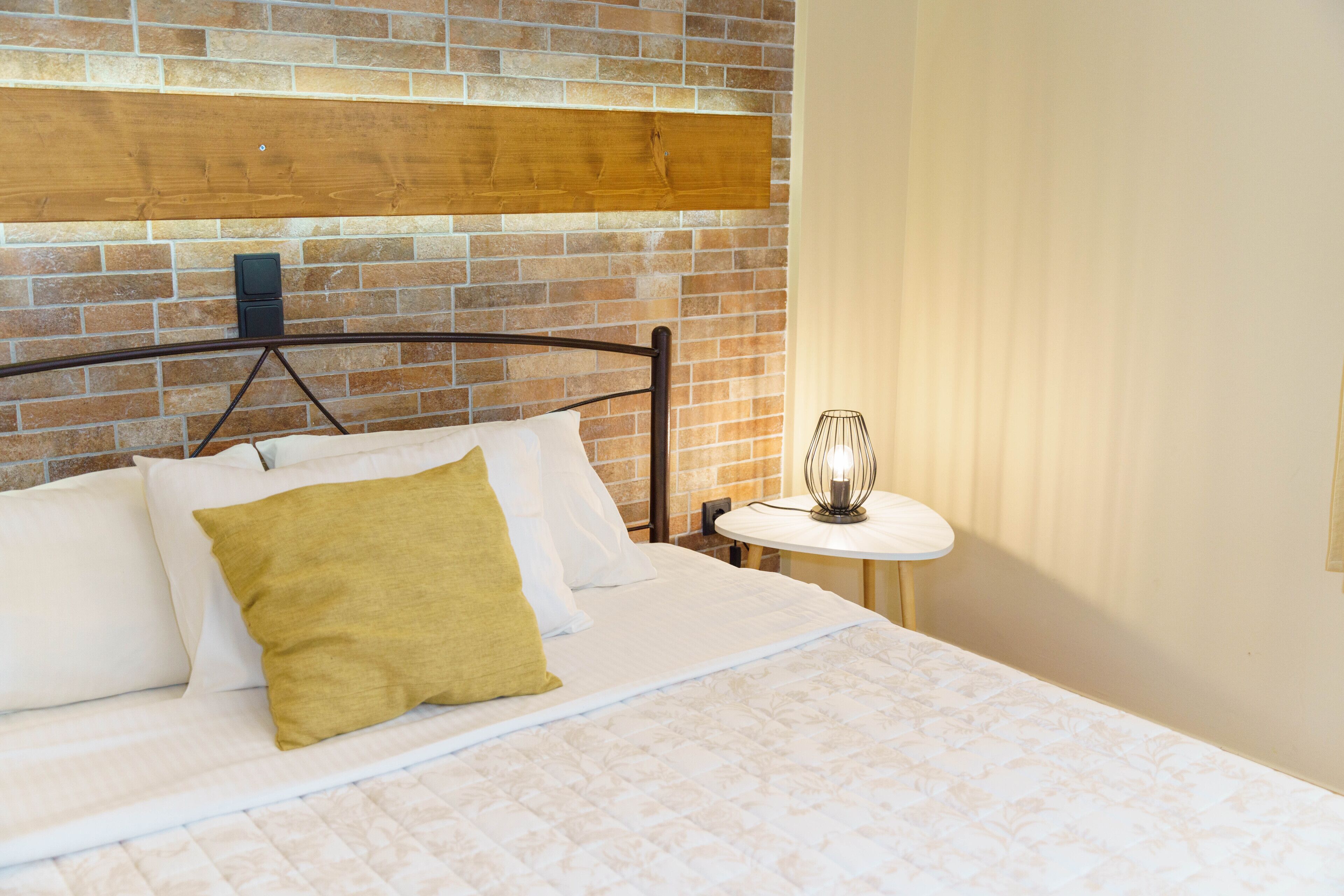 A neatly made bed with white and mustard pillows, a wooden headboard, and a small table with a lamp beside it.