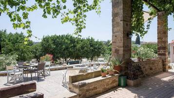 An outdoor seating area with stone benches and tables, surrounded by greenery and a clear sky.