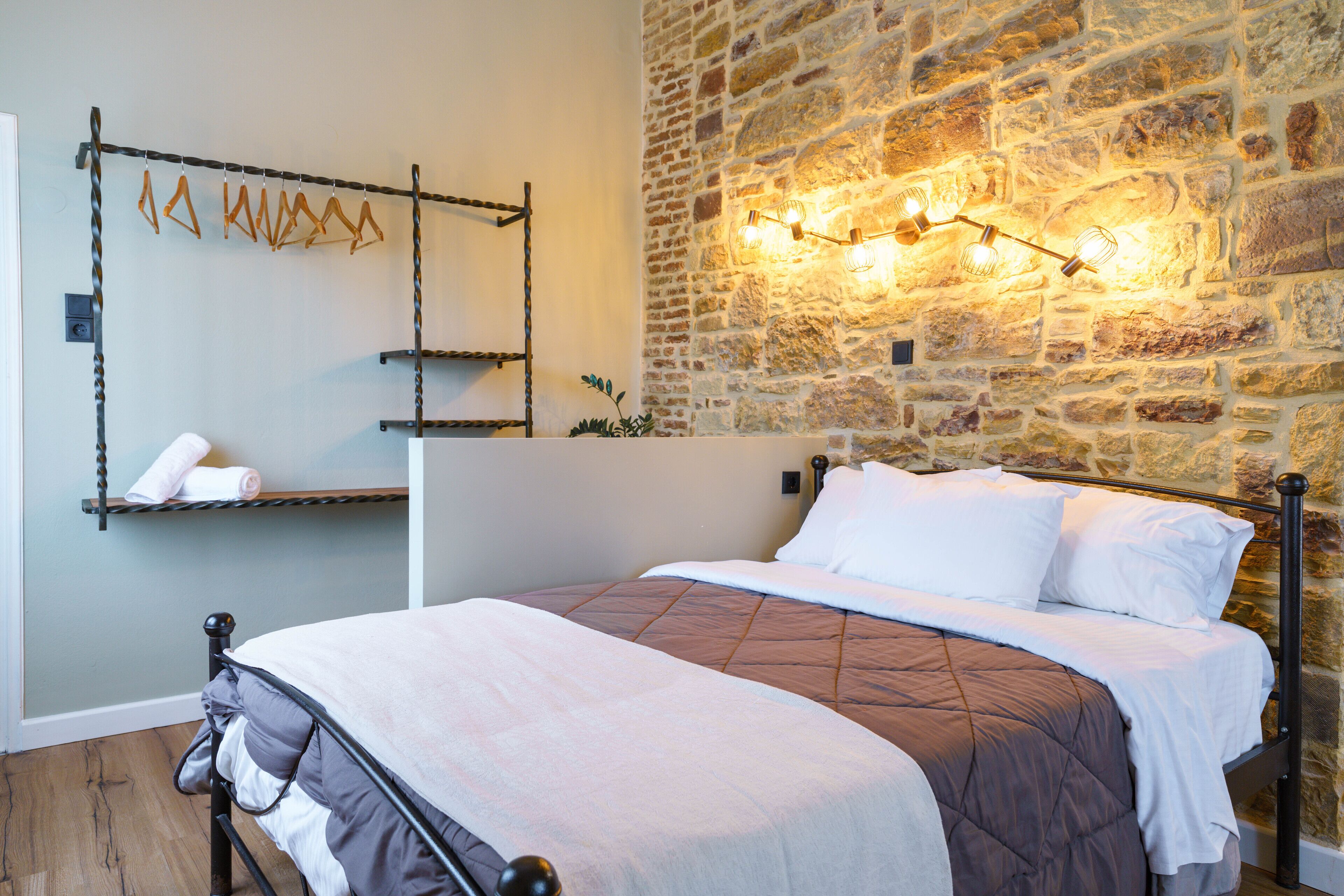 Um quarto com parede de pedra, cama com colcha marrom, cabideiro com toalhas e luminária fixada na parede.