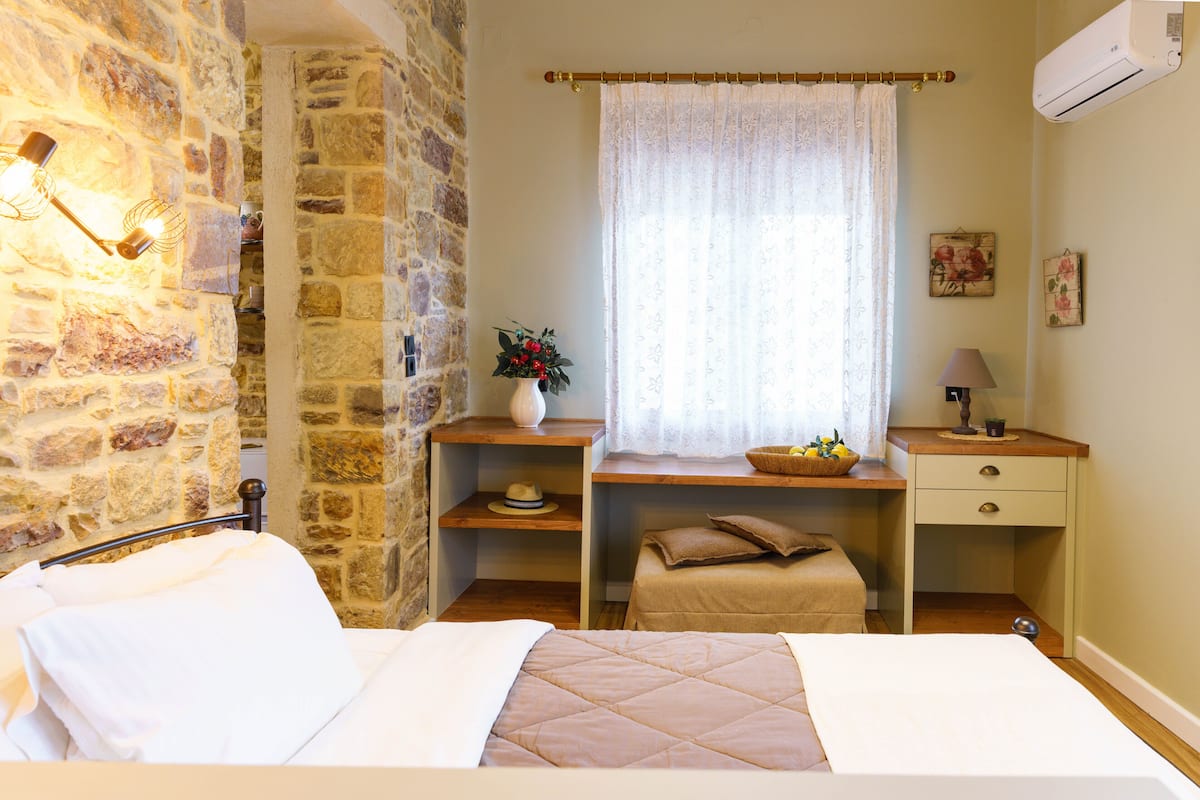 Un dormitorio con pared de piedra, una cama con ropa de cama blanca, un escritorio de madera con un florero y una ventana con cortina translúcida.
