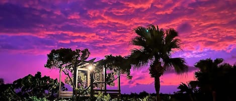Una estructura tipo quiosco rodeada de abundante follaje y palmeras bajo un cielo de atardecer intenso.