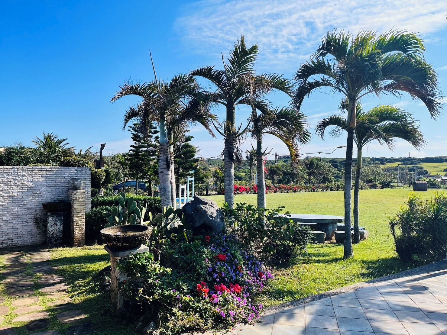 A garden with palm trees, a stone wall, and a stone fountain.