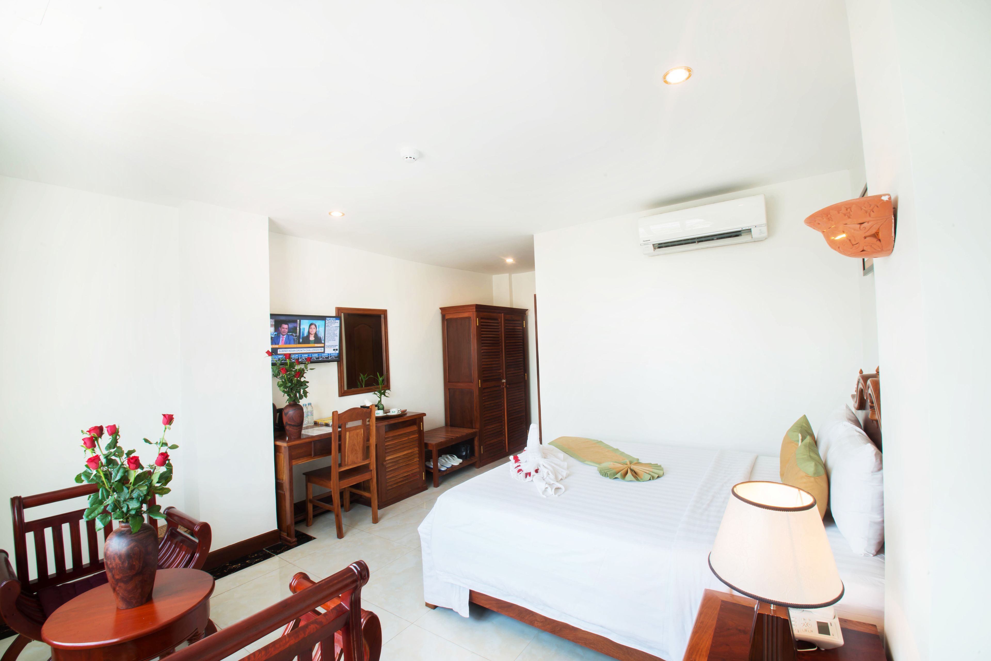 Una habitación de hotel con cama, escritorio, silla, mesa con un jarrón de flores y un equipo de aire acondicionado.
