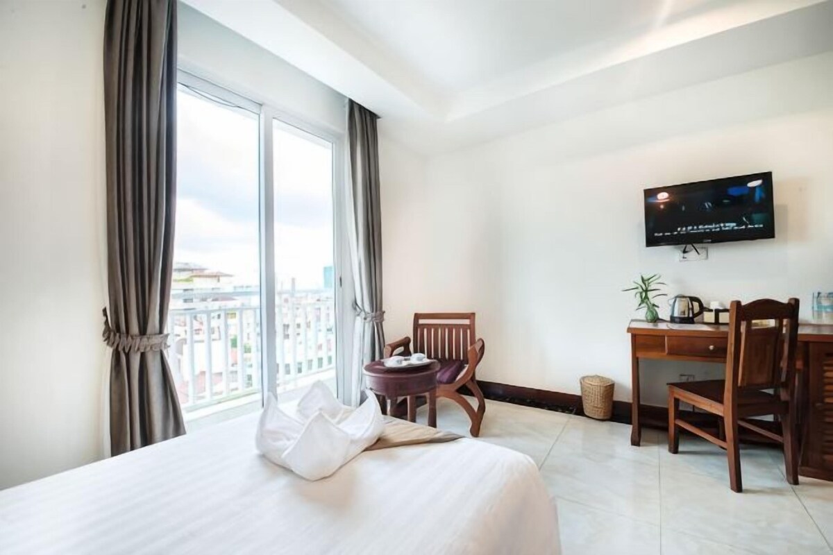 Quarto de hotel com uma cama, uma escrivaninha, uma cadeira, uma televisão e vista para a cidade através de amplas janelas.