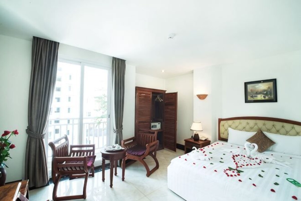 Een hotelkamer met een bed, een bureau, een stoel, een raam met gordijnen, een nachtkastje en een wandlamp.