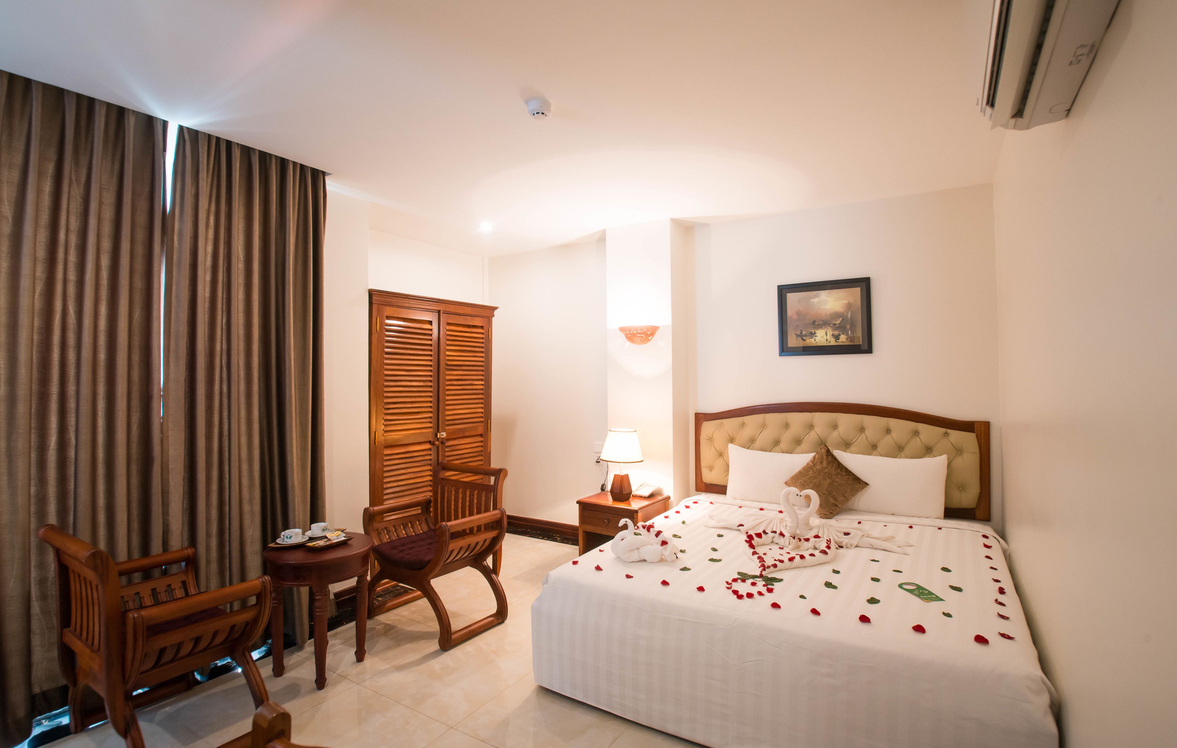A hotel room with a bed, a wooden chair, a small table, a lamp, and a picture on the wall.