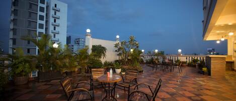 A rooftop terrace with a checkered floor, several tables and chairs, potted plants, and a view of the city skyline at dusk.