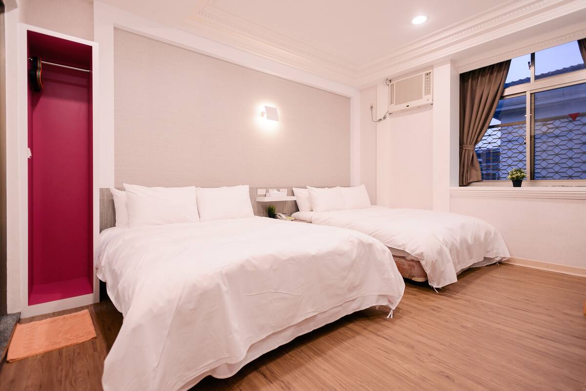 Hotelkamer met twee bedden, een rode deur, een raam met gordijnen en een luchtverkoeler aan de muur.