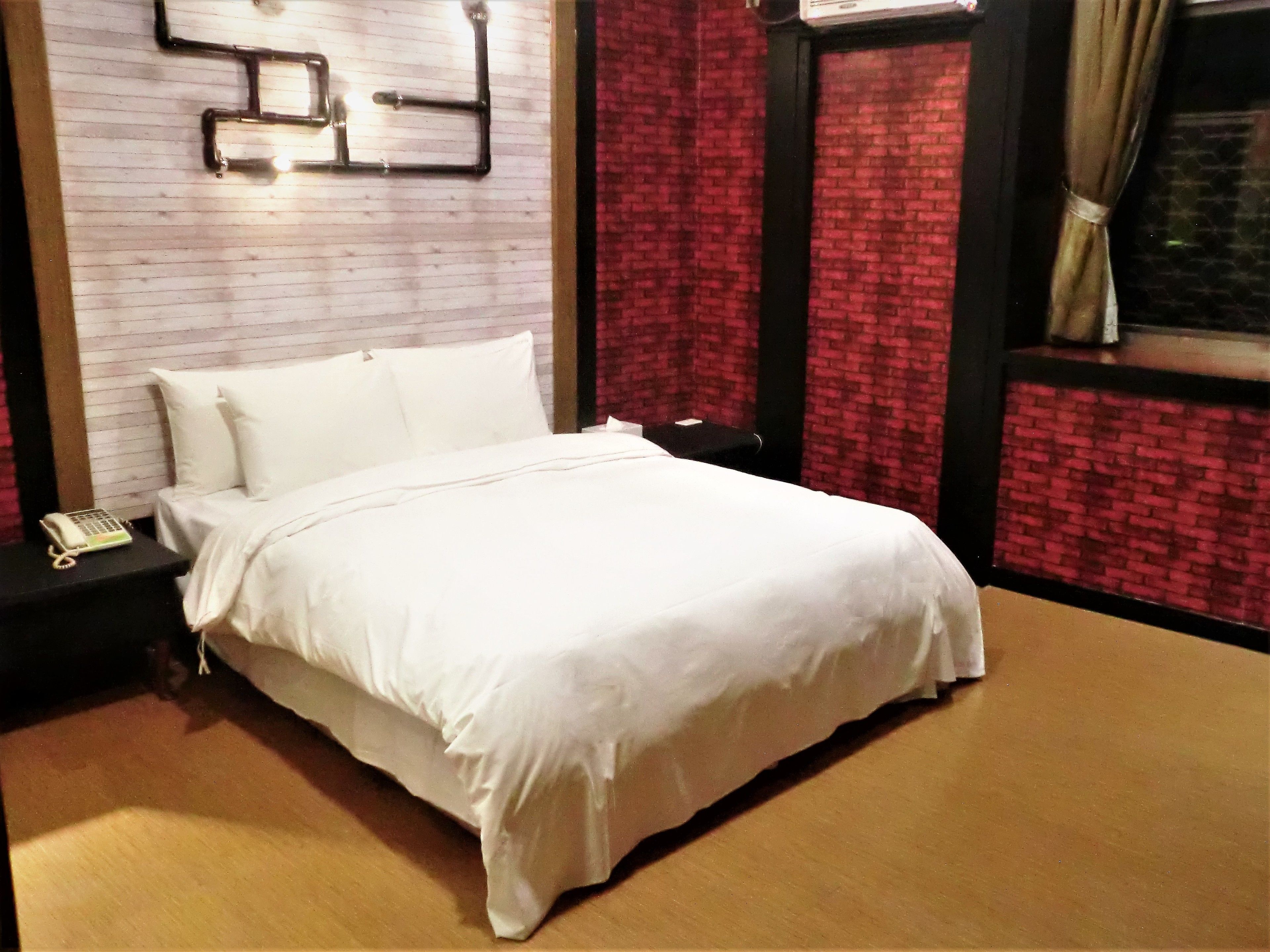 Una cama bien hecha en una habitación de hotel, con una pared de ladrillo visto, un teléfono en la mesita de noche y una ventana con cortinas.