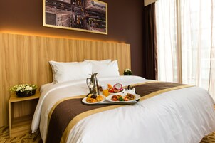 A hotel room with a neatly made bed, a tray with breakfast items, a cityscape painting, and a nightstand with a flower.
