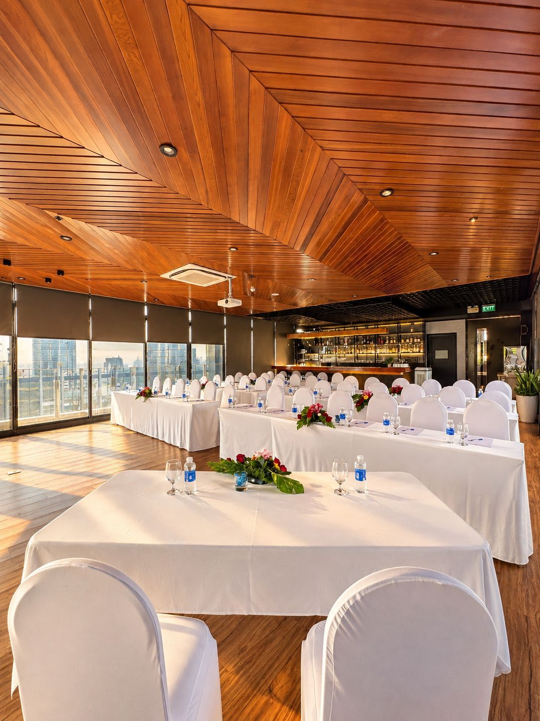 Un salón de conferencias con techo alto, mesas y sillas blancas, una zona de bar y ventanas amplias con vistas a la ciudad.