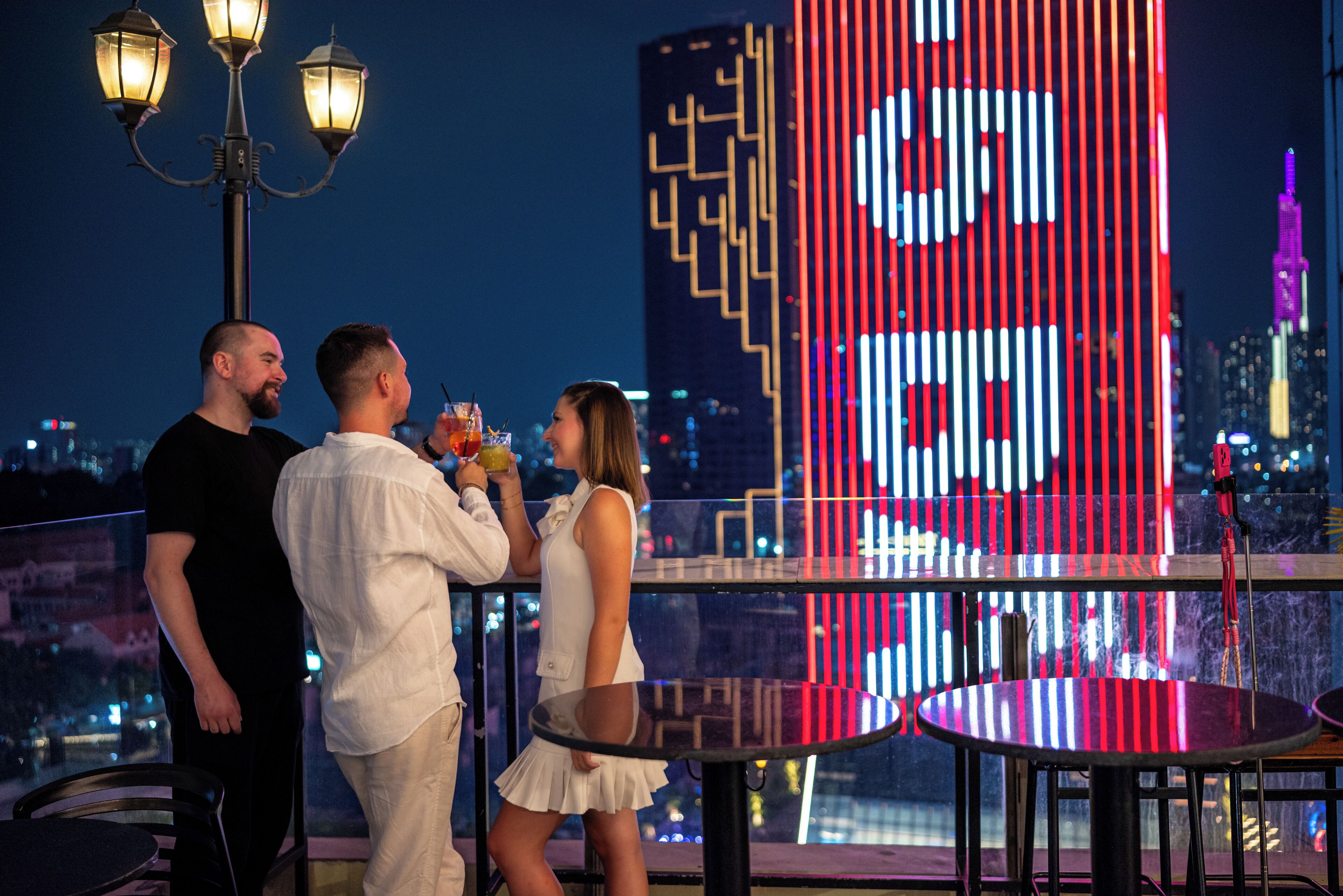 Un bar situé sur un toit, offrant une vue panoramique sur la ville, avec un grand écran numérique affichant un logo, et quelques personnes qui savourent des boissons.