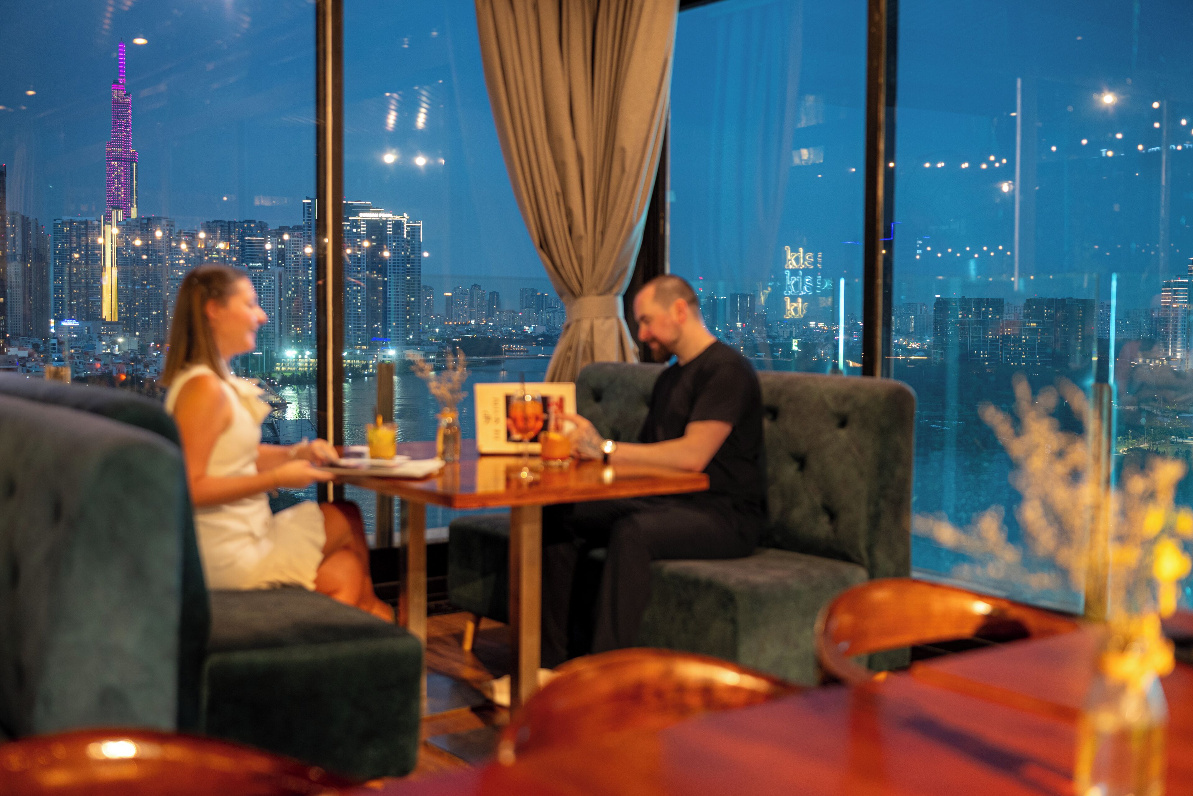 Un couple est assis à une table, avec une vue sur la ville, un rideau et un vase rempli de fleurs.