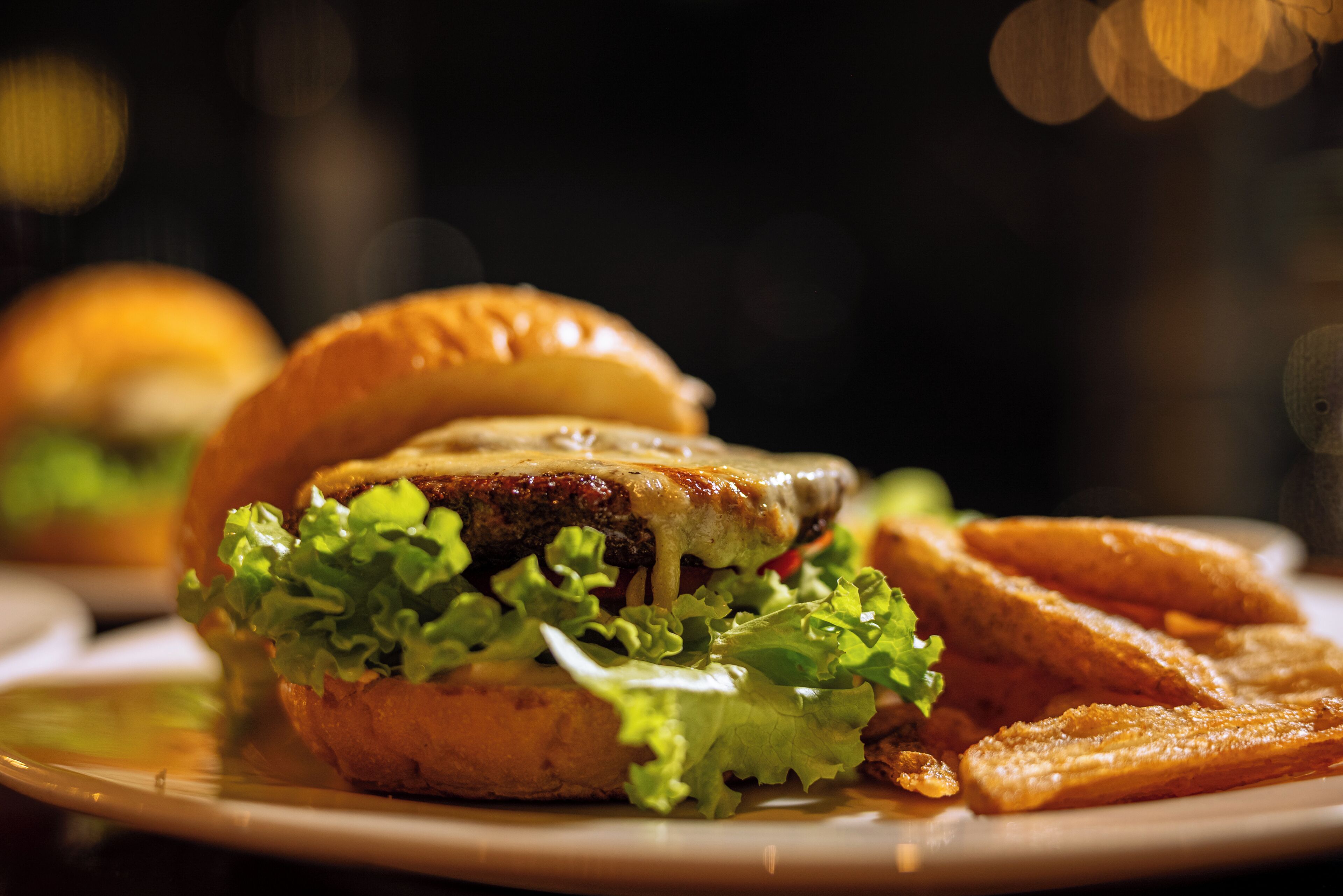 Primer plano de una hamburguesa con carne, queso y lechuga en un pan, acompañada de papas fritas en un plato.