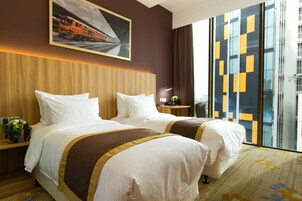 A hotel room with two beds, a wooden headboard, a large window with a view of a building, and a painting of a train on the wall.