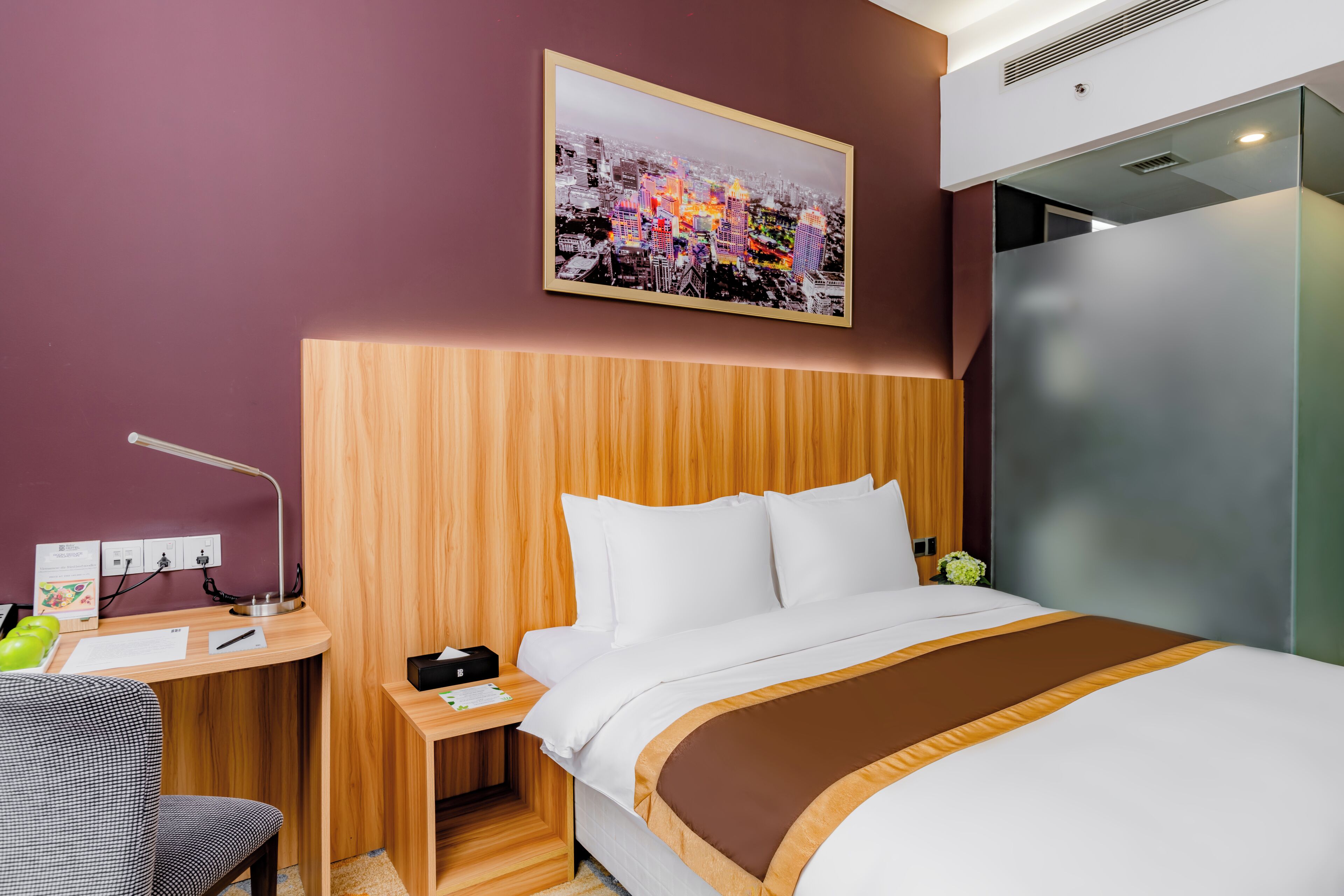 A hotel room with a bed, a desk with a lamp, a nightstand, and a wall-mounted picture.