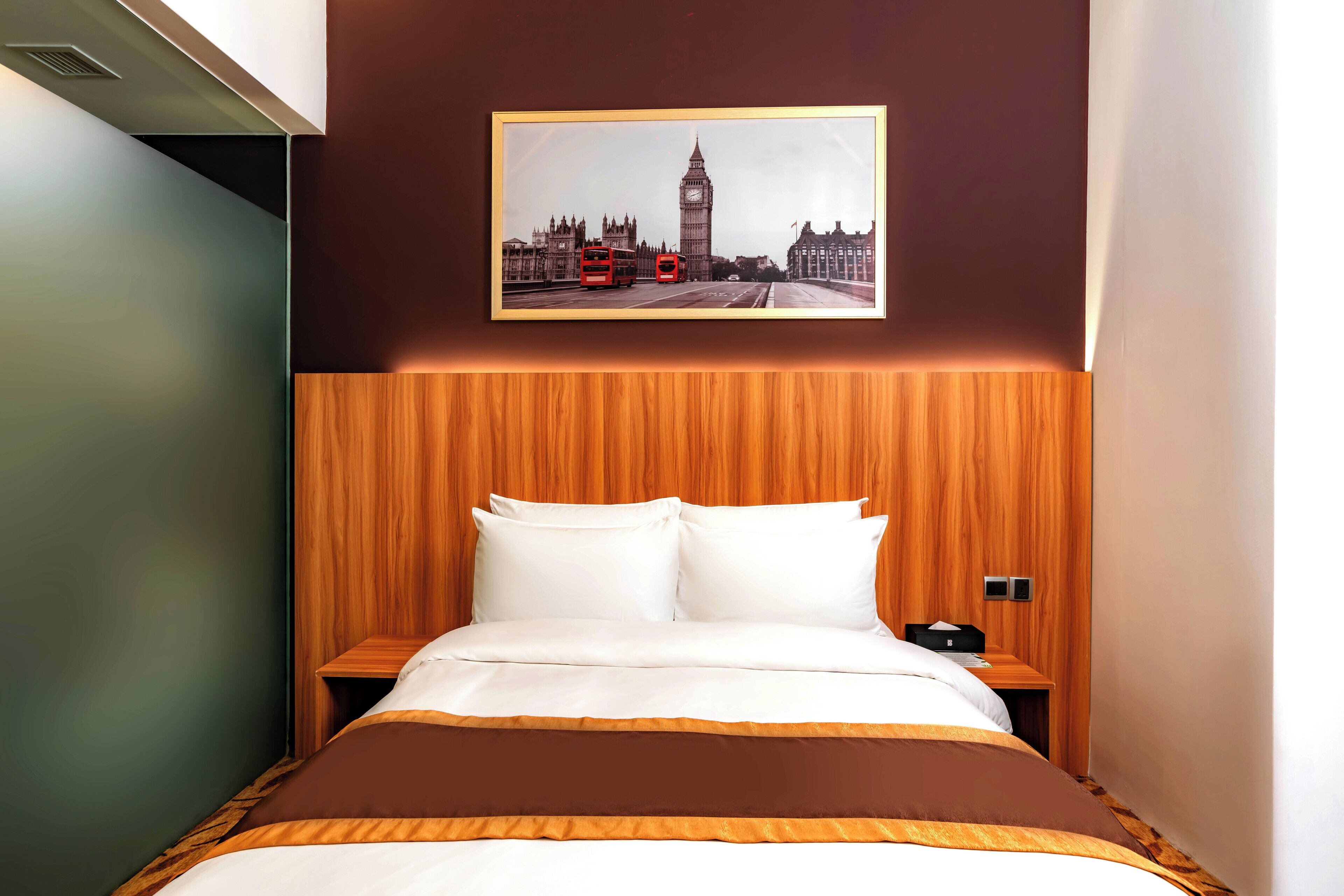 Une chambre d’hôtel avec un lit, une tête de lit en bois, une table de chevet et un tableau représentant Londres accroché au mur.