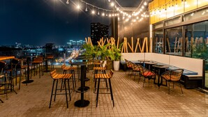 An outdoor terrace with tables and chairs, string lights, and a view of the city at night. - Bay Hotel Ho Chi Minh (Ho Chi Minh City)