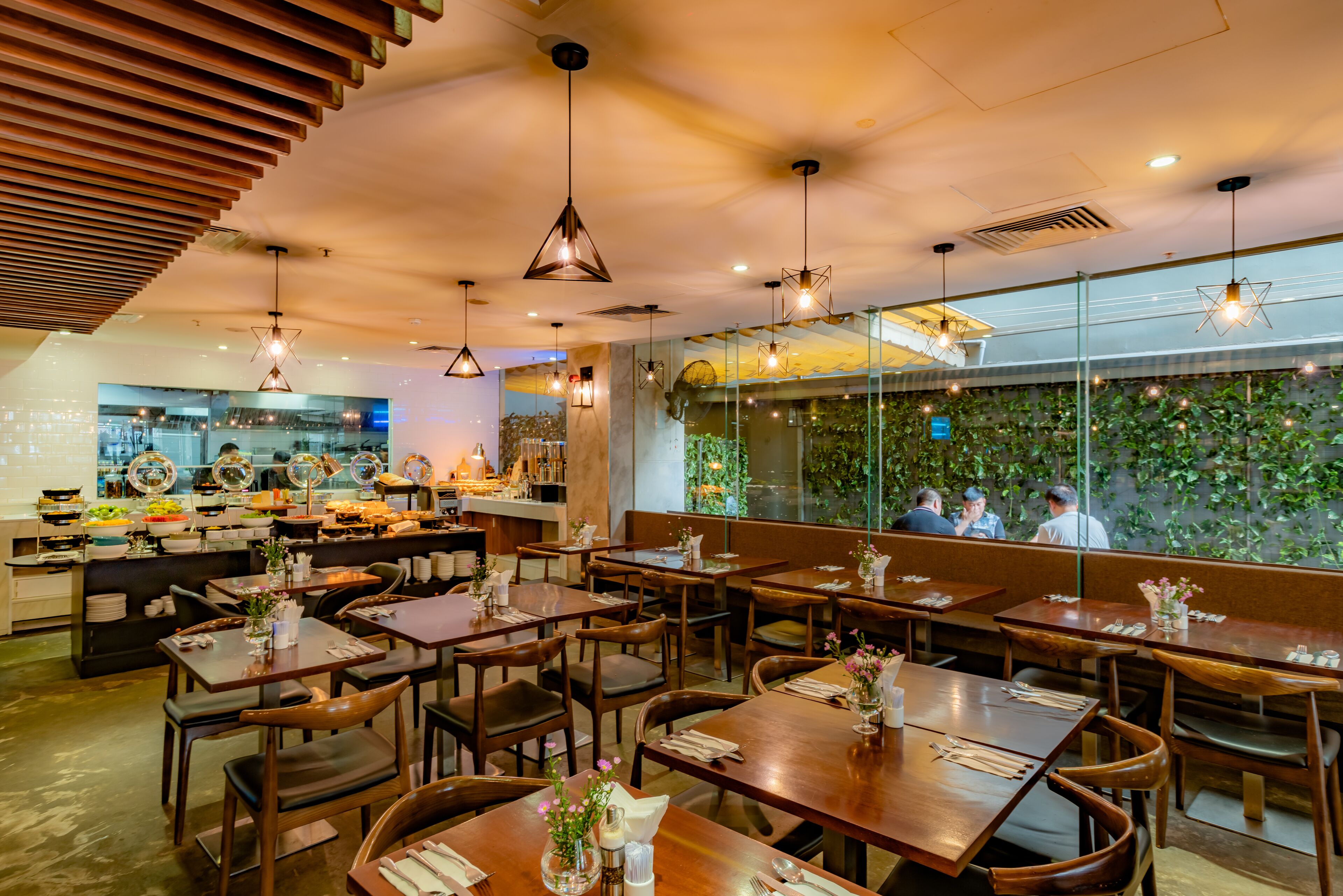 Interior de restaurante con mesas y sillas de madera, lámparas colgantes y vistas a un jardín a través de un muro de cristal.