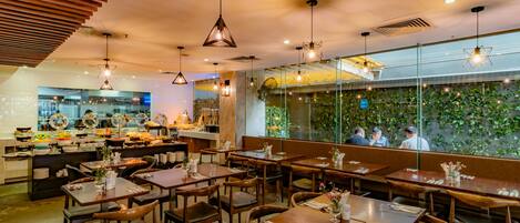Interior de restaurante con mesas y sillas de madera, lámparas colgantes y vistas a un jardín a través de un muro de cristal.