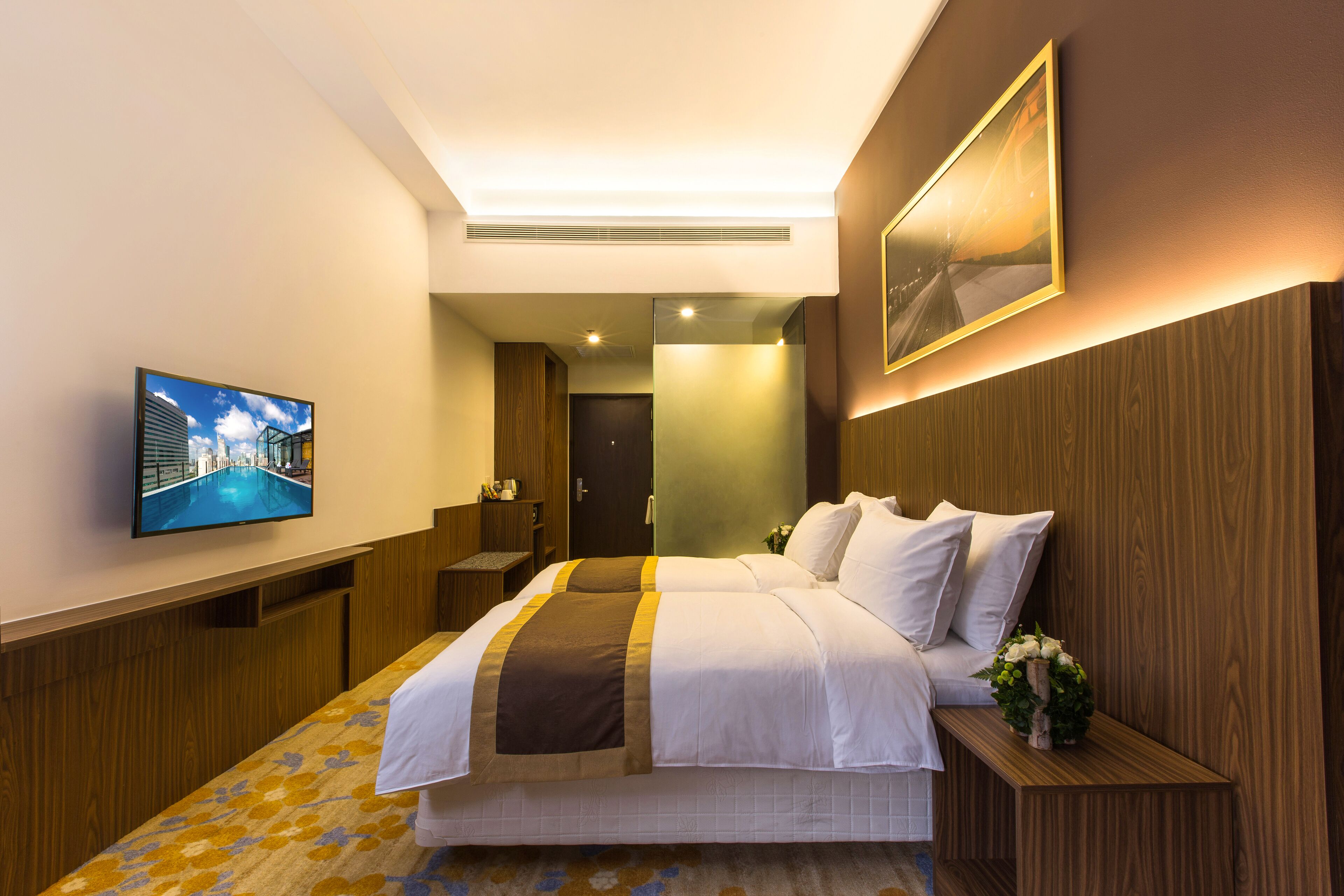 A hotel room with a large bed, a television on the wall, a wooden headboard, and a small table with a vase of flowers.