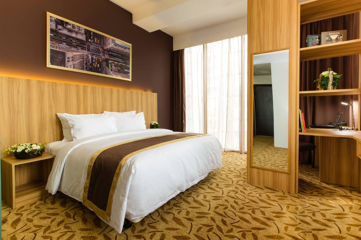 Ein Hotelzimmer mit einem großen Bett, einem Holzkopfende, einem Schreibtisch mit Stuhl, einem Bücherregal und einem großen Fenster mit Vorhängen.