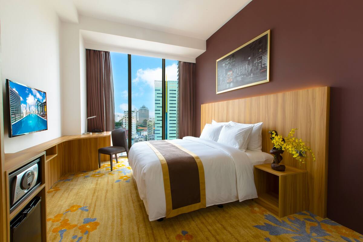 Ein Hotelzimmer mit einem großen Bett, einem Schreibtisch, einem Sessel, einem Fernseher und Blick auf die Stadt durch große Fenster.