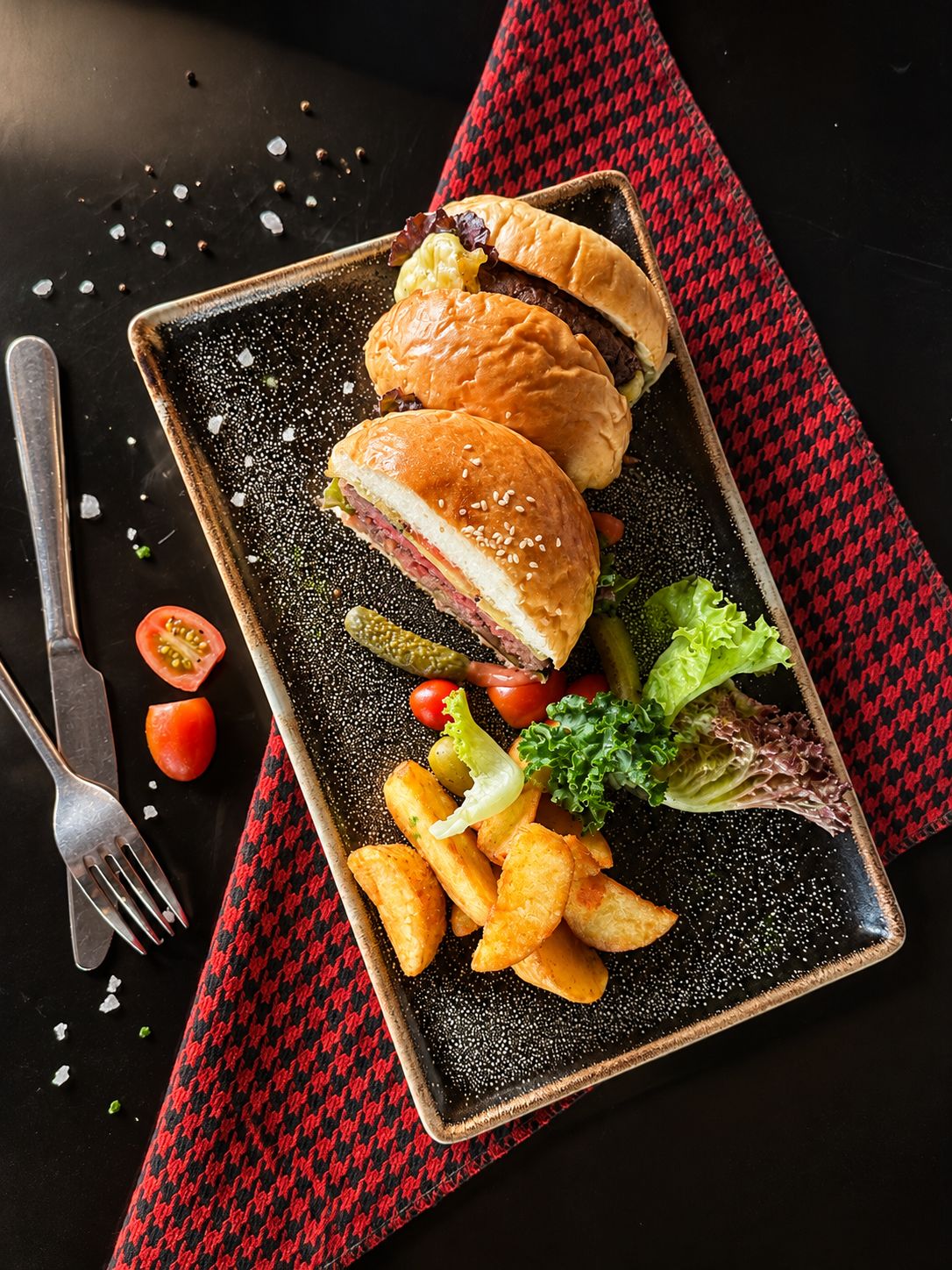 Comida que incluye un sándwich, papas fritas y pepinillos sobre un plato negro con una servilleta de cuadros rojos y negros debajo.