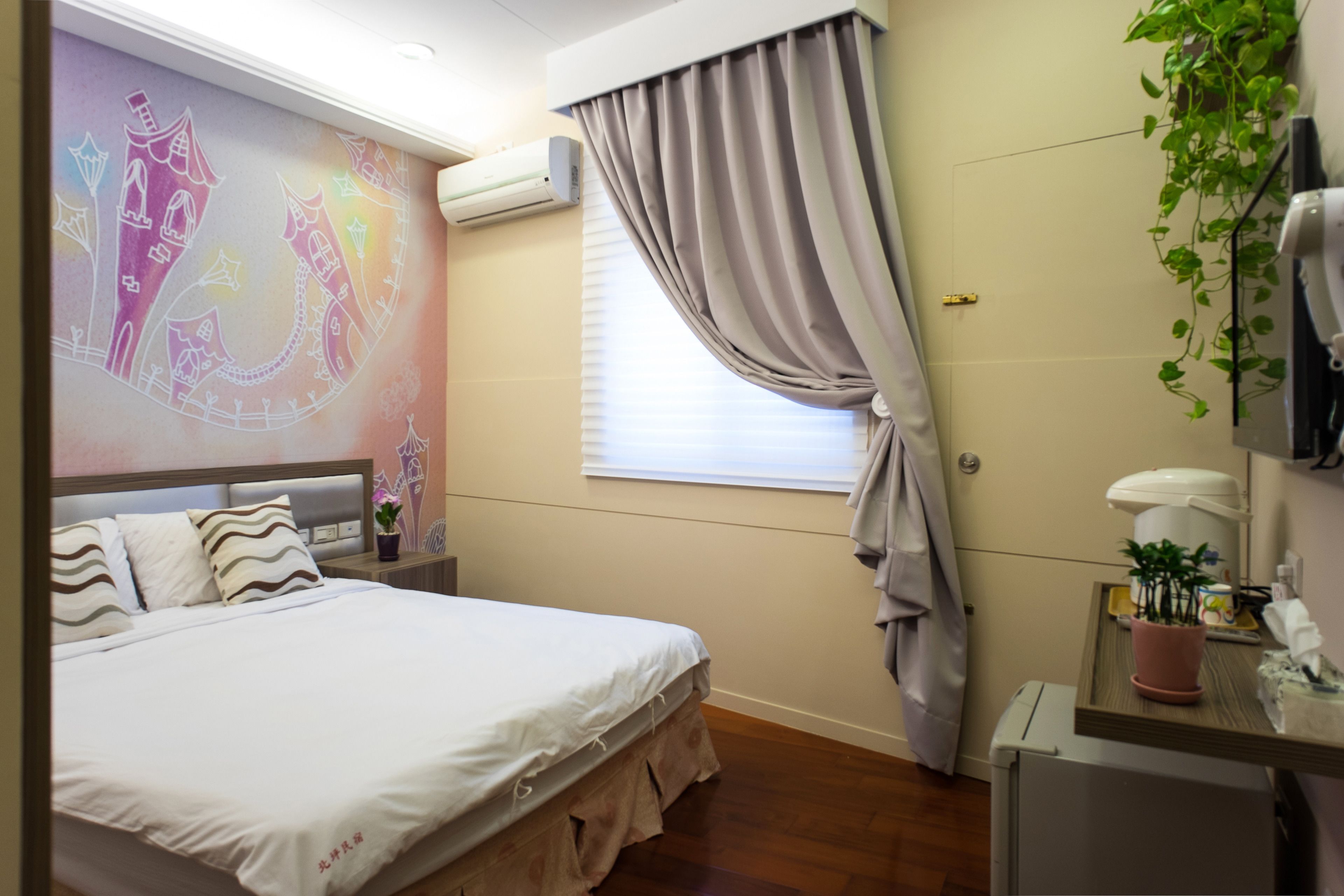A bedroom with a bed, a wall mural, an air conditioning unit, a window with curtains, a bedside table with a plant, and a small desk with a lamp.
