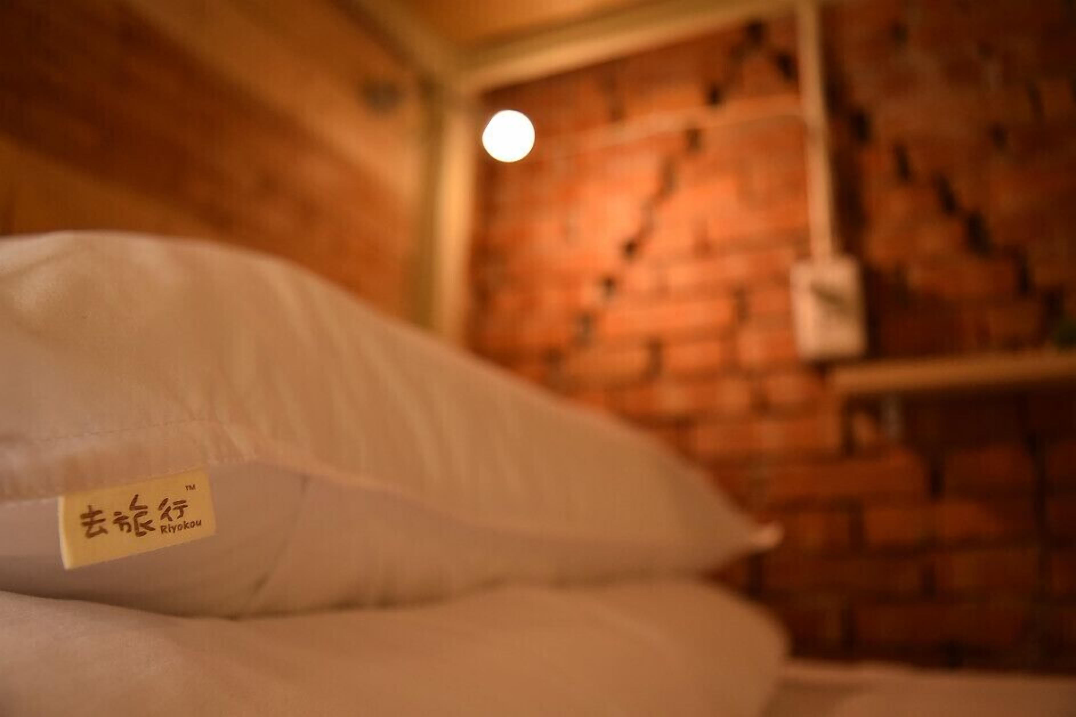 A bed with a pillow labeled "Ryokou" in a room with a brick wall and a hanging light fixture.