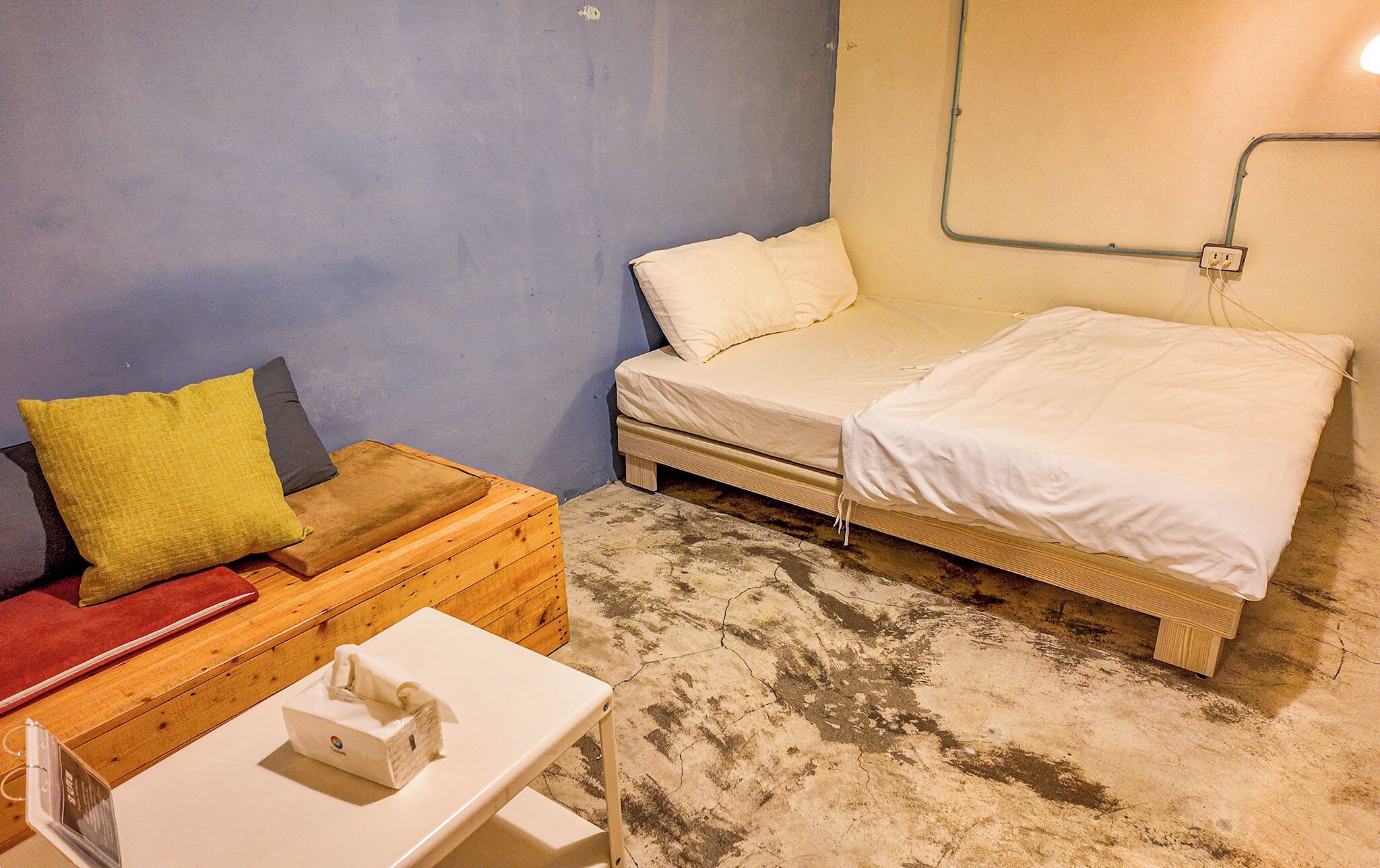 A small, compact room with a bed, a wooden bench with cushions, and a small table with a tissue box.