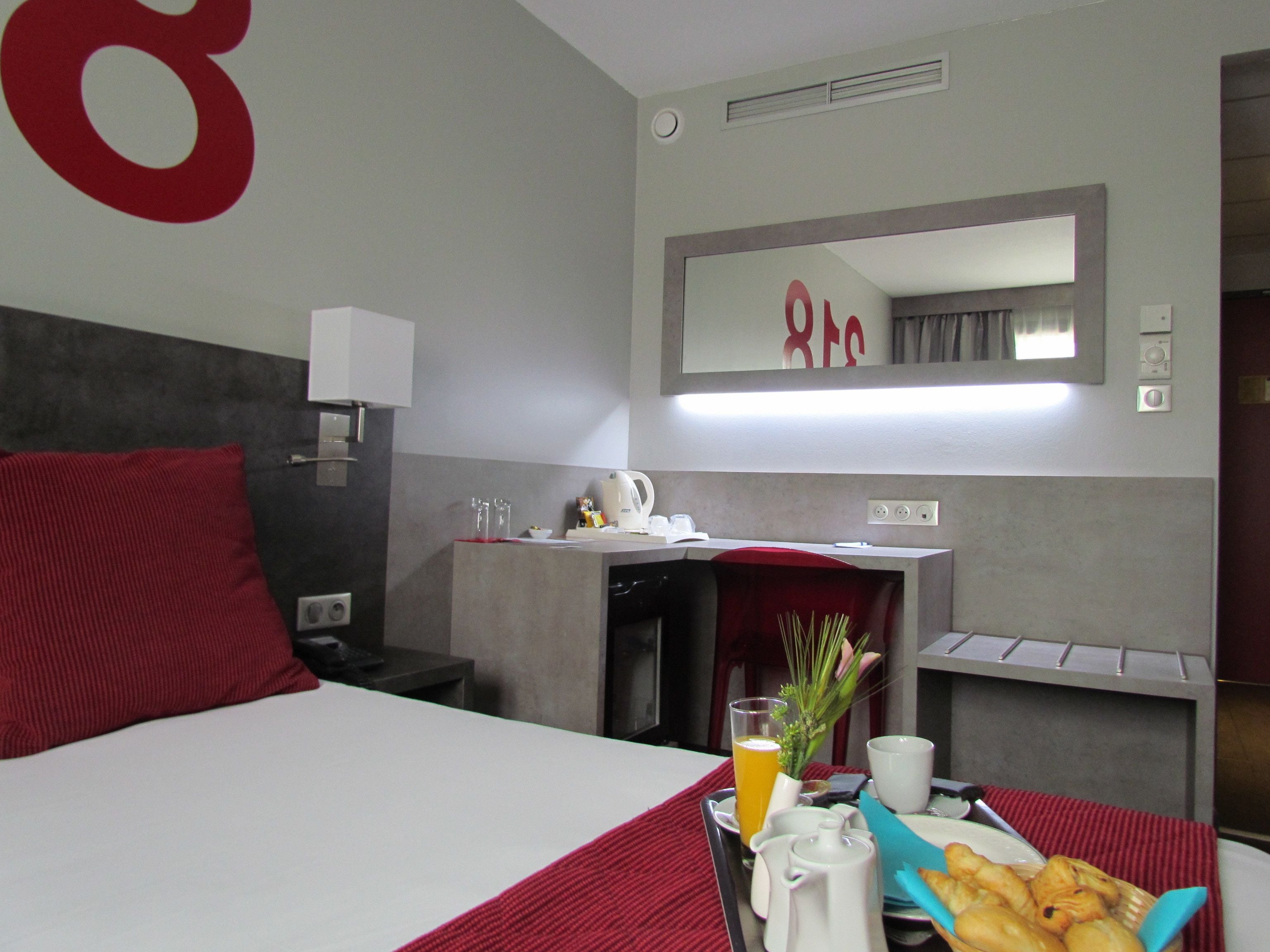 A hotel room with a bed, a desk, a mirror, and a breakfast tray with orange juice, a cup, and pastries.