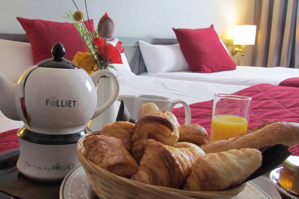 Una habitación de hotel con una cama, almohadas, un hervidor de agua, una cesta de cruasanes, un vaso de jugo de naranja y una bandeja.