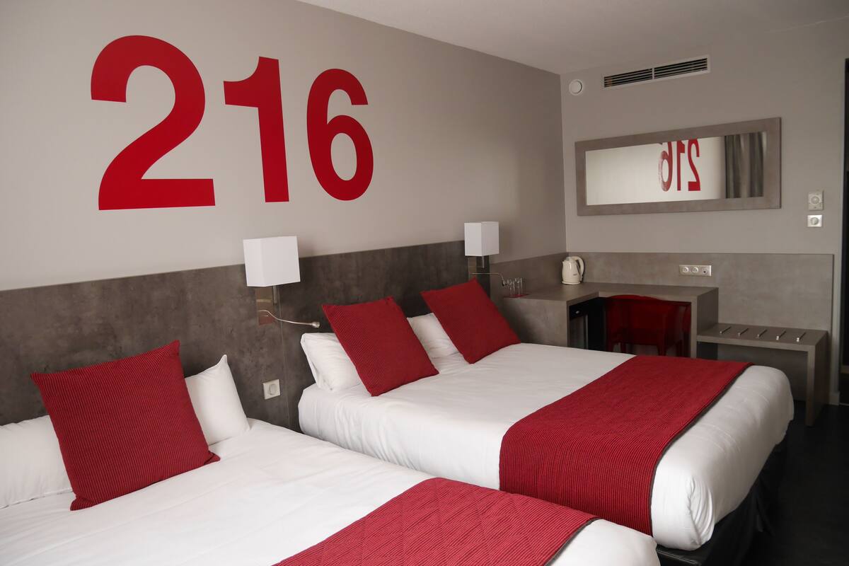 Habitación de hotel con dos camas, cada una con ropa de cama roja y blanca, un gran número '216' en la pared, un escritorio con una silla y un espejo sobre el escritorio.