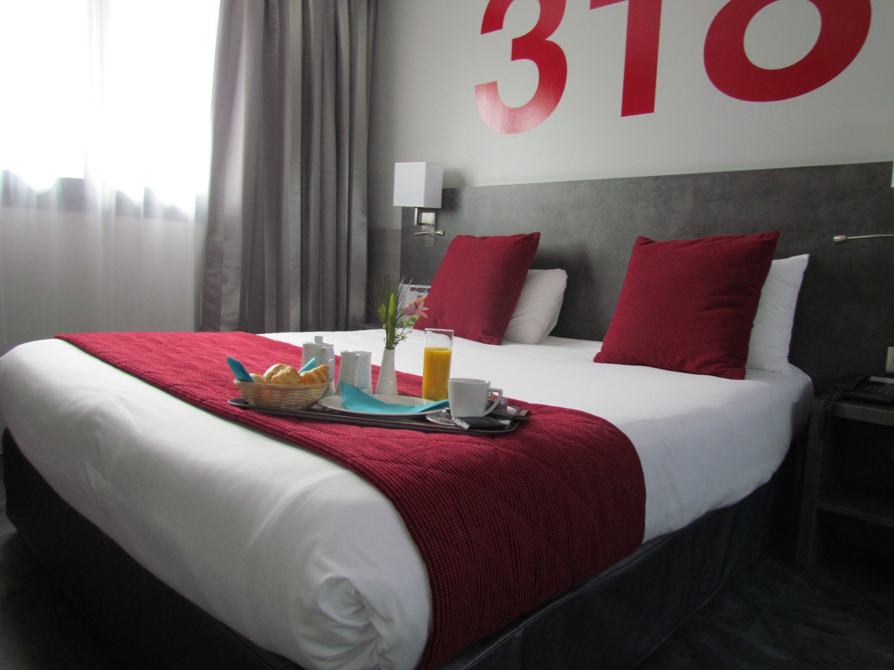 A hotel room with a large bed, red and white bedding, a breakfast tray with croissants, a vase with flowers, and a large number '310' on the wall.