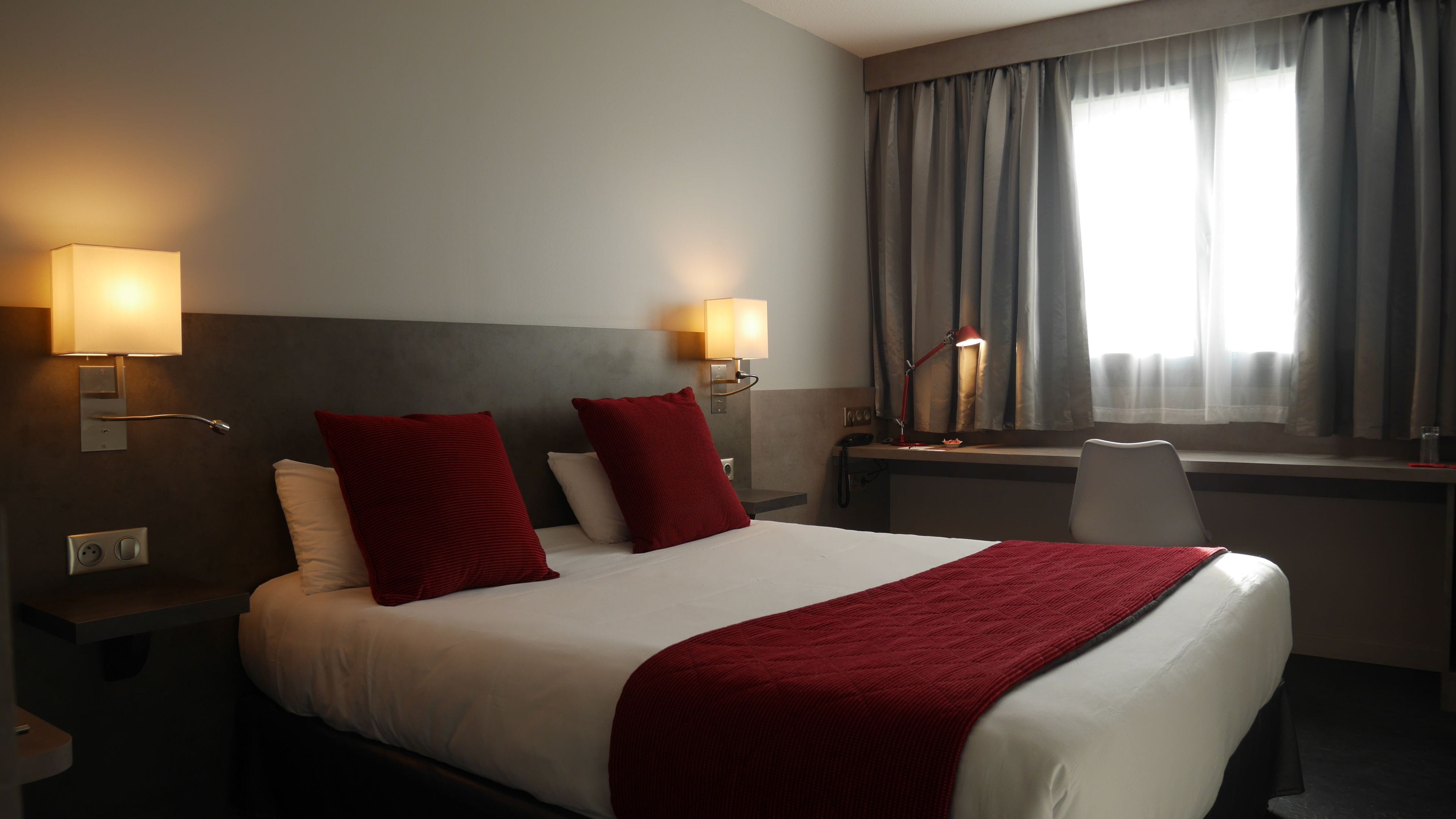 Habitación de hotel con una cama grande, dos almohadas rojas, un escritorio con lámpara y una ventana con cortinas.