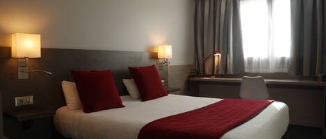 A hotel room with a large bed, two red pillows, a desk with a lamp, and a window with curtains.