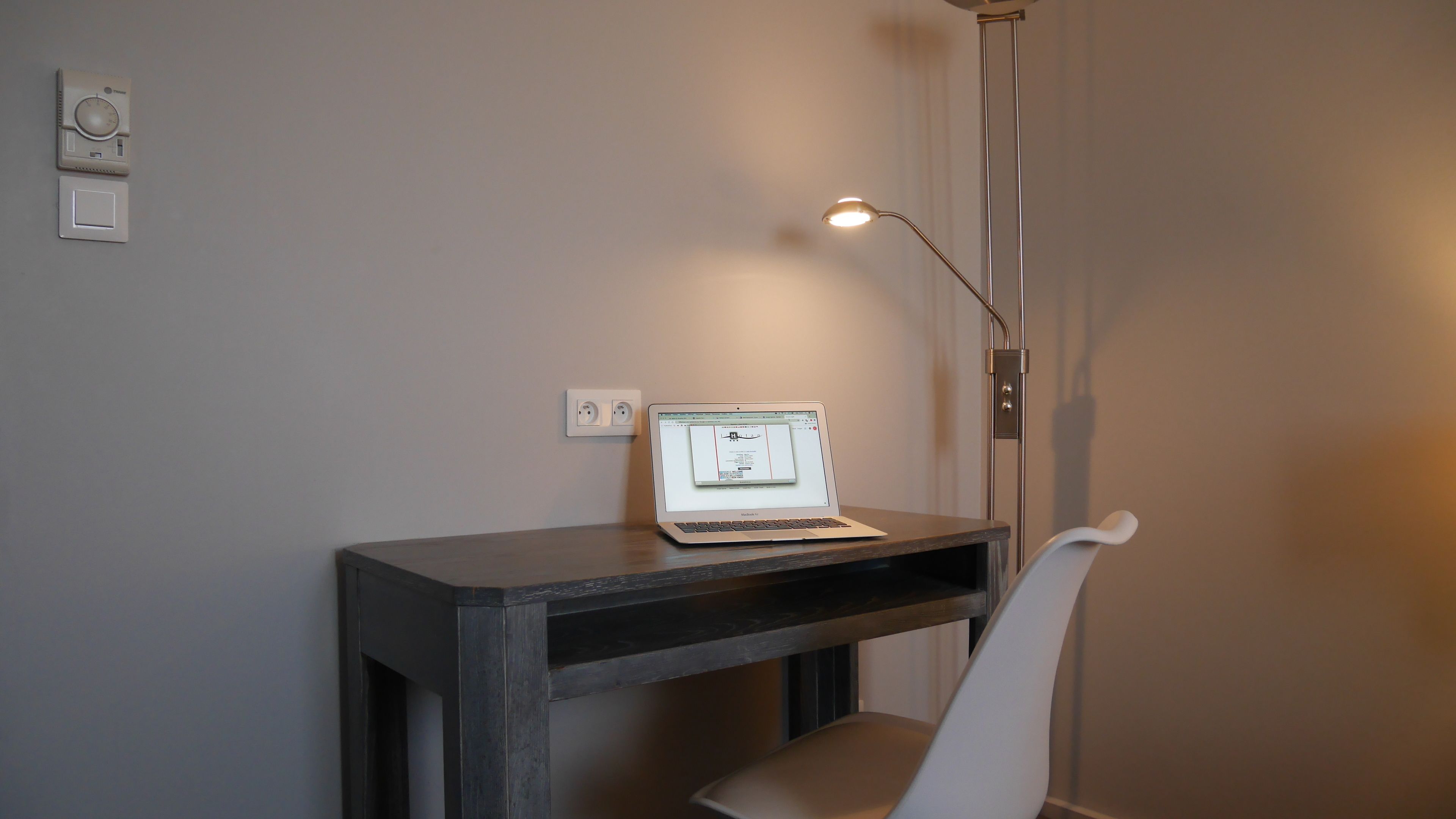 A laptop on a dark wooden desk with a white chair, a floor lamp, and a wall socket.