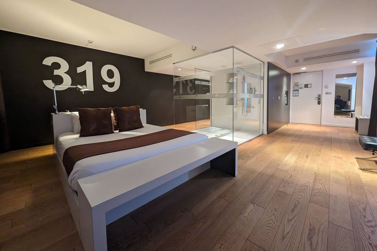 Una habitación de hotel moderna con una cama grande, una ducha con cerramiento de vidrio y un cartel con número en la pared.