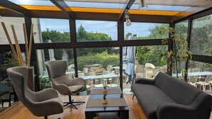 A modern interior with a wooden floor, two armchairs, a sofa, and a coffee table. Large windows offer a view of an outdoor patio area with tables and chairs. - Hôtel L'Hotan (Portet-sur-Garonne)