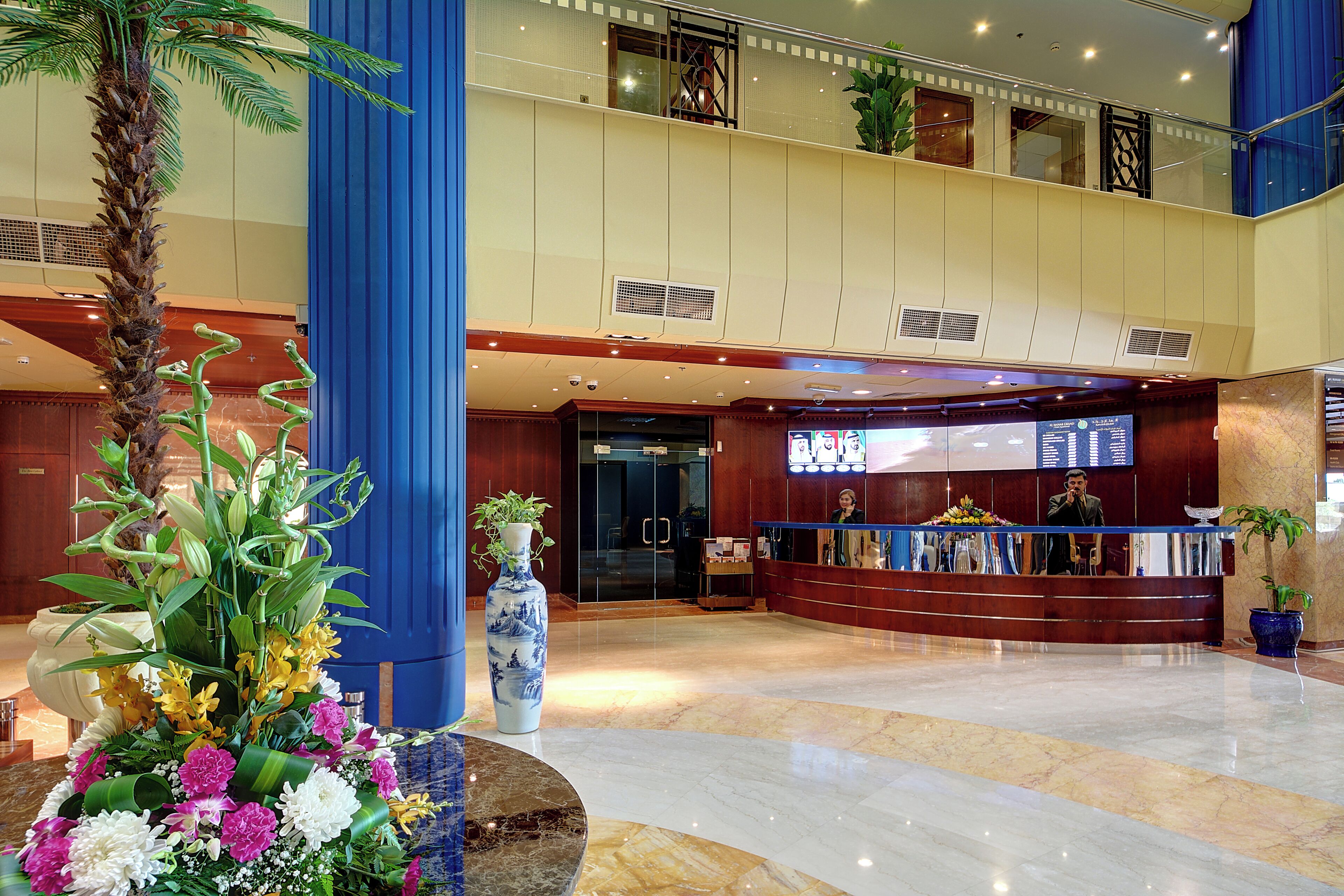 A hotel lobby with a reception desk, a large vase with flowers, and a palm tree.