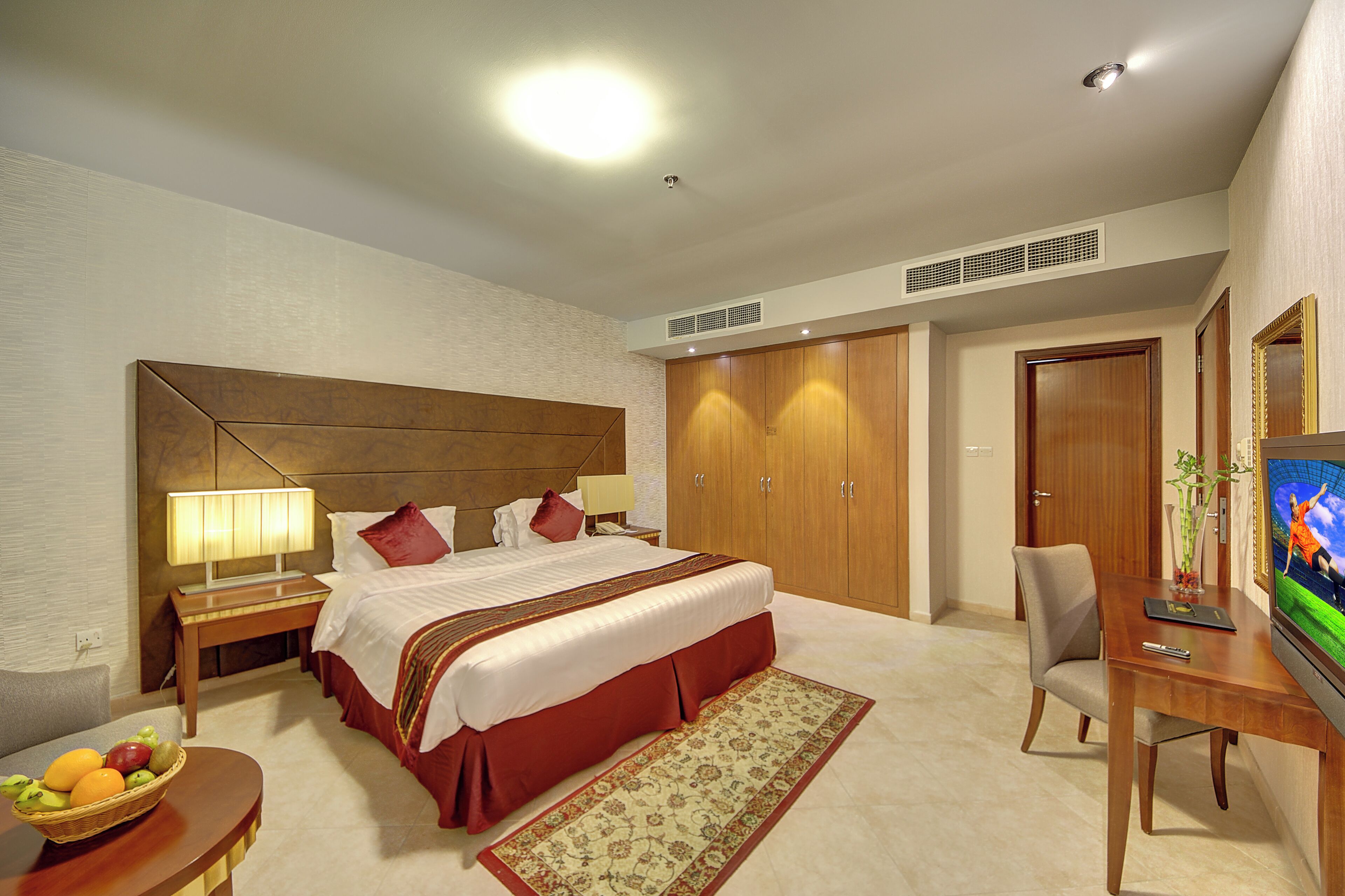 Une chambre d’hôtel comprenant un lit, un bureau avec une télévision, une chaise, une table de chevet, une lampe, un panier de fruits et une armoire.