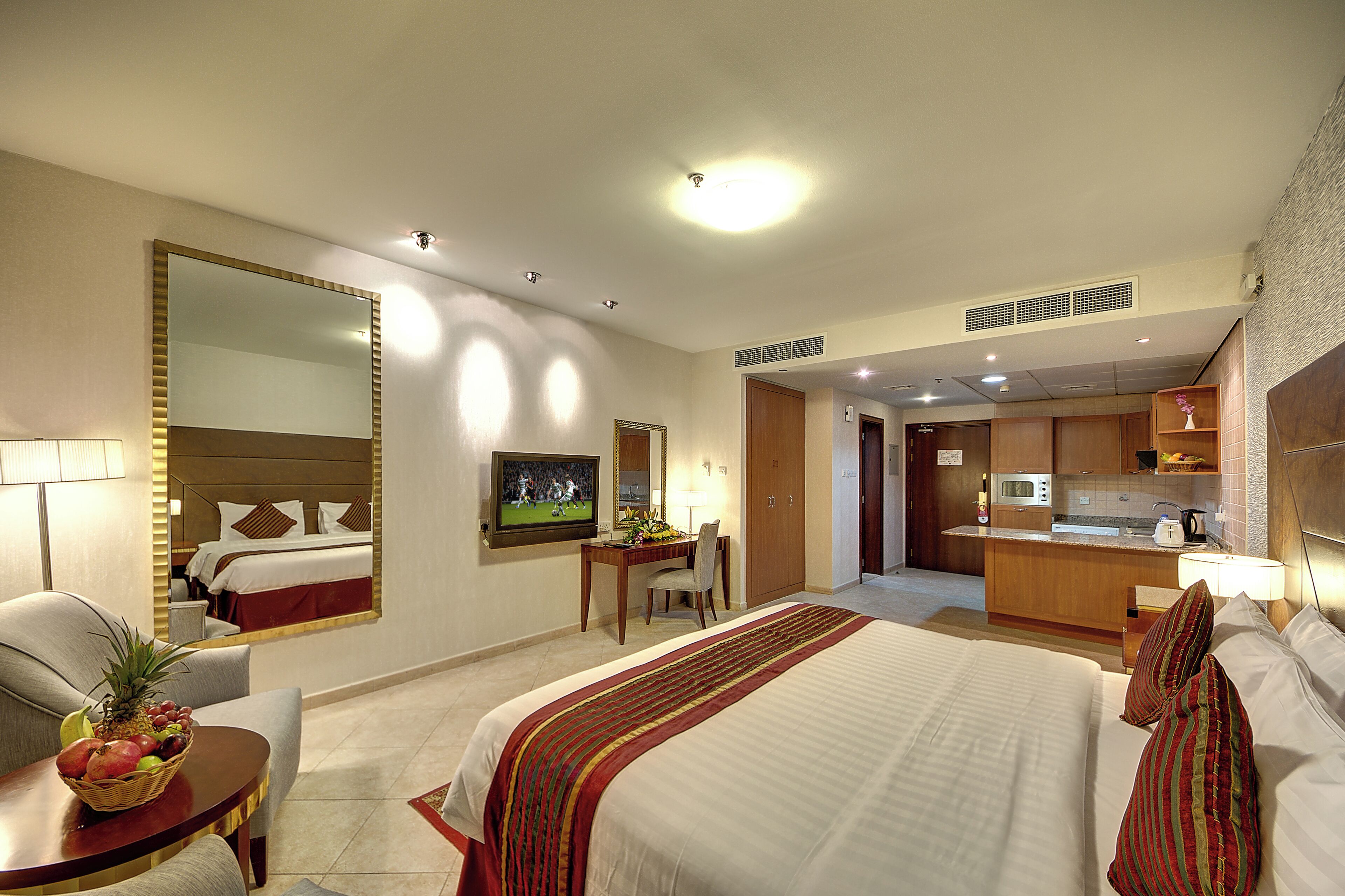 A hotel room with a large bed, a desk, a chair, a television, and a kitchenette.