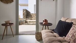 A modern living room with a beige sofa, a dark blue pillow, and a woven basket. There are two small round tables with bowls and a vase. A woven lamp hangs on the wall, and a stone wall is visible through the glass door. - Ianemi Suites by K4 Kythnos (Kithnos)