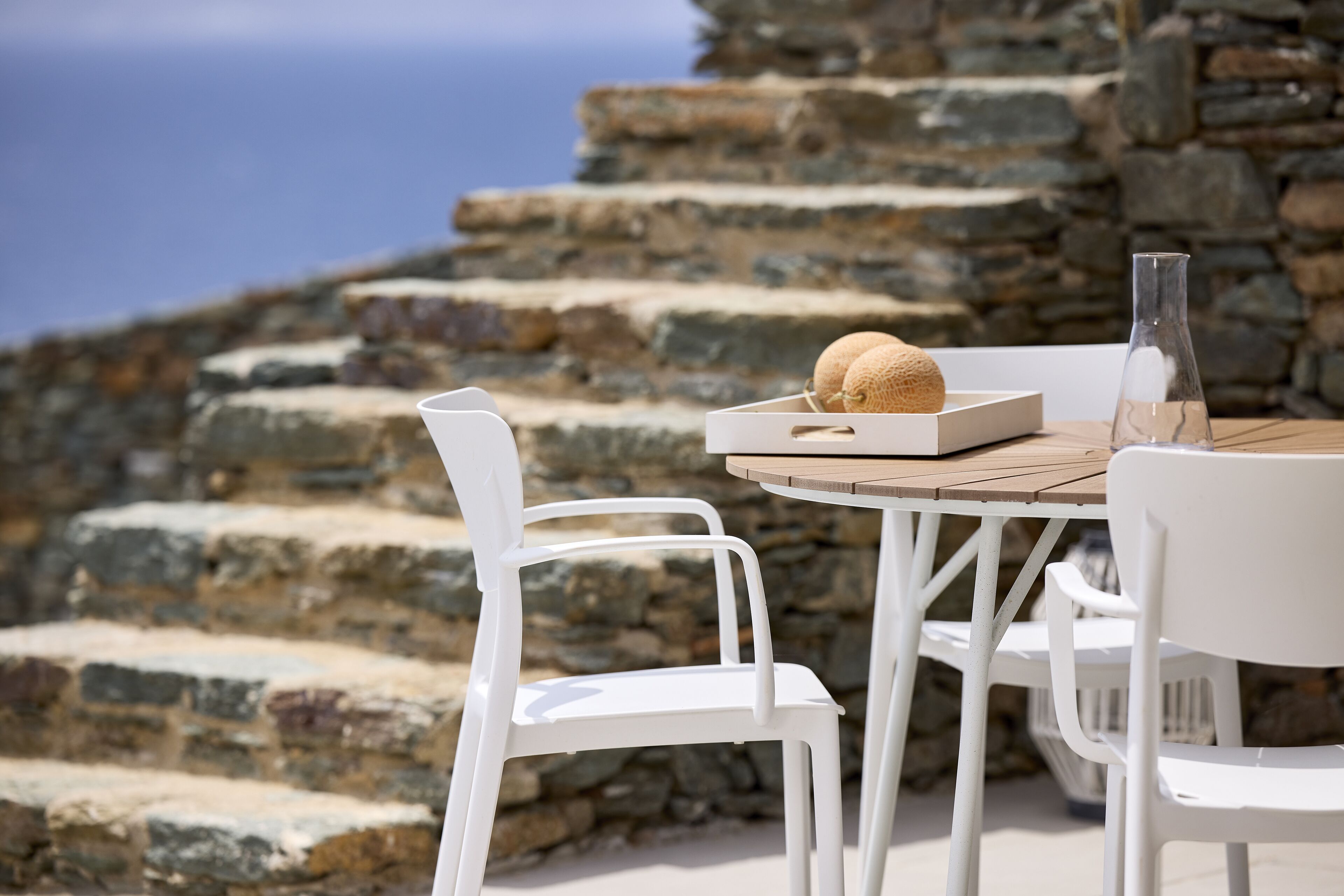 Une table avec un plateau en bois, un verre et une chaise blanche, le tout placé devant un mur de pierre avec des marches menant à un point de vue.