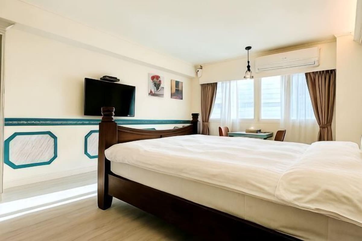 A hotel room with a bed, a television, a window with curtains, and a wall-mounted air conditioner.
