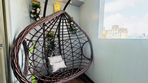 Una silla colgante de mimbre con cojines, piso de madera, ventana con vista a edificios e iluminación fijada a la pared.