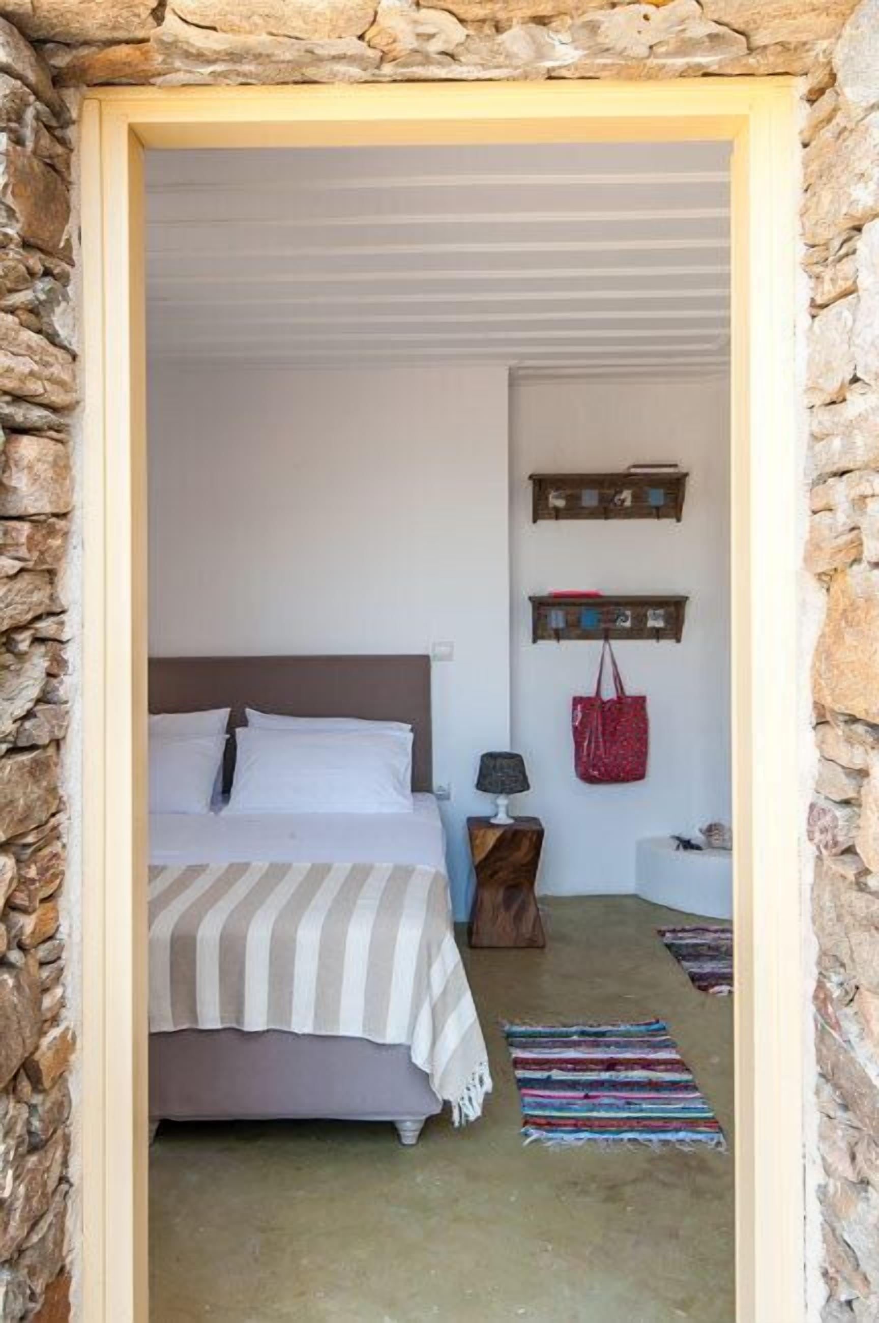 A bedroom with a bed, a nightstand, a lamp, a red bag, and a wall-mounted shelf.
