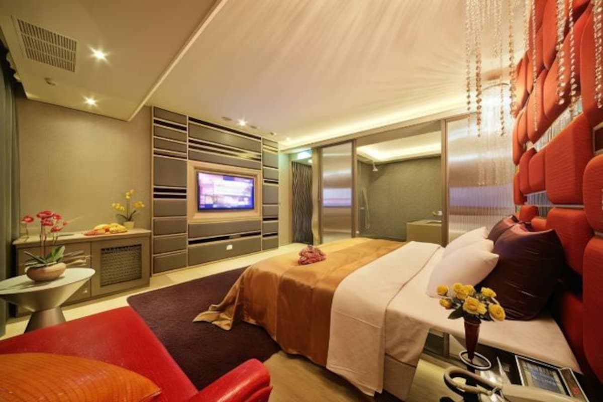 A modern hotel room with a large bed, a television, a seating area with a red sofa, and a small table with flowers.